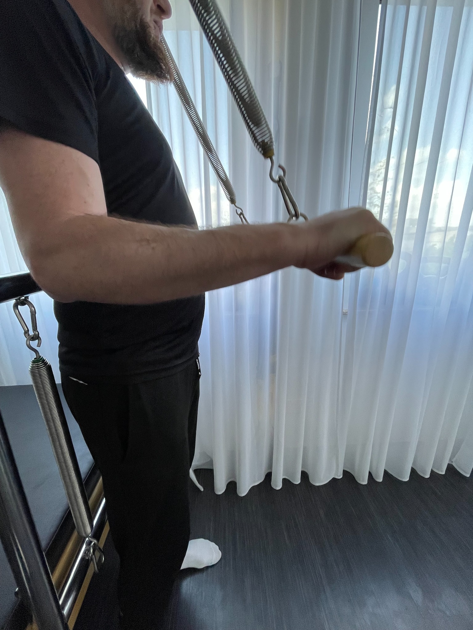 Mężczyzna ćwiczy na reformerze Pilates z użyciem sprężyn, widoczne białe zasłony w tle.