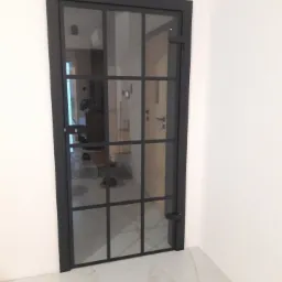 Nowoczesne drzwi metalowe z szybami w czarnej ramie, widok na wnętrze mieszkania z białymi ścianami i marmurową podłogą.