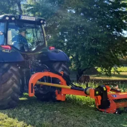 Traktor z pomarańczową kosiarką rotacyjną przy pracy na trawniku, widoczny operator w kabinie, drzewa w tle.