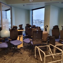 Space Job s.c - Puste krzesła biurowe i elementy wyposażenia ustawione w pomieszczeniu biurowym z widokiem na miasto za oknem, prawdopodobnie przygotowane do sprzątania lub przeprowadzki.