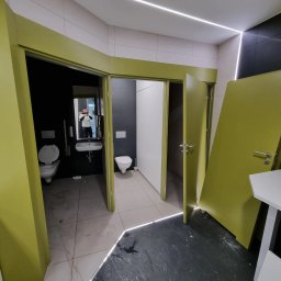 Space Job s.c - Wnętrze biurowej toalety z dwoma kabinami, widoczne dwie muszle klozetowe, umywalka z lustrem, czarne ściany i podłoga, oświetlenie LED na suficie, drzwi w kolorze limonki, odbicie fotografa...