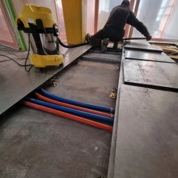 Space Job s.c - Instalacja podłogi podniesionej w trakcie remontu, widoczne rury instalacyjne, odkurzacz przemysłowy i pracownik układający płyty.