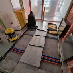 Space Job s.c - Montaż podłogi podniesionej w biurze: mężczyzna w czarnym ubraniu instaluje metalowe płyty podłogowe, widoczne rury w kolorze niebieskim i czerwonym, żółty odkurzacz przemysłowy obok.