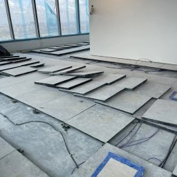 Space Job s.c - Montaż podłogi technicznej w biurze: płyty podłogowe ułożone częściowo, widoczne przewody i elementy konstrukcyjne, w tle okno z widokiem na miasto.