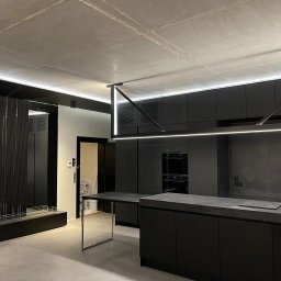 Space Job s.c - Nowoczesna, minimalistyczna kuchnia w odcieniach szarości i czerni z surowym betonowym sufitem, oświetleniem LED i wyspą kuchenną z blatem imitującym kamień.