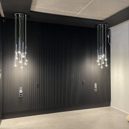 Space Job s.c - Ciemna, pionowo żłobkowana ściana z dwoma wiszącymi lampami sufitowymi z wieloma punktami świetlnymi, widoczne gniazdka elektryczne.