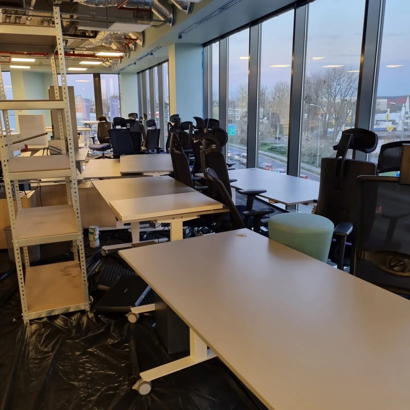 Przygotowanie biura do malowania: biurka i krzesła zabezpieczone folią, metalowy regał z półkami, widok na miasto za oknem.