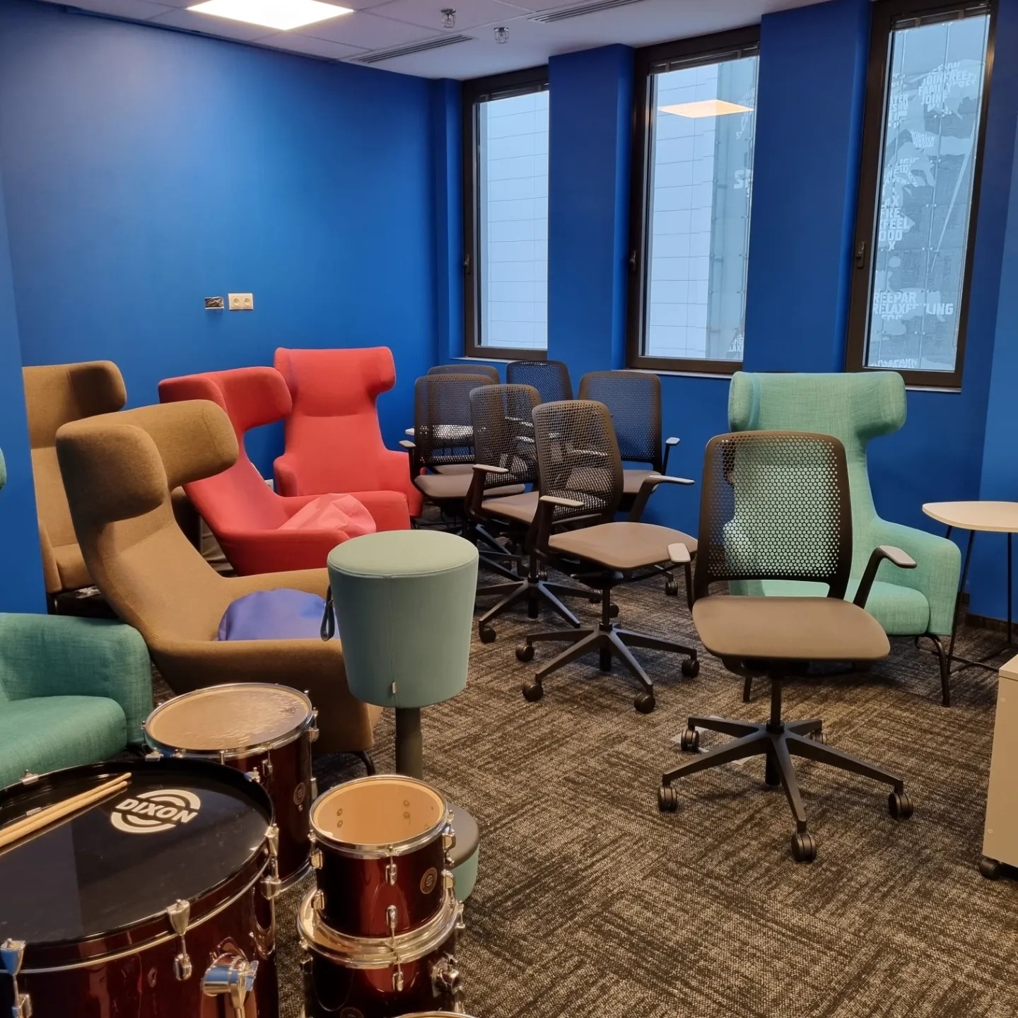 Biuro z niebieskimi ścianami, różnokolorowymi fotelami, biurowymi krzesłami i zestawem perkusyjnym Dixon na dywanie.