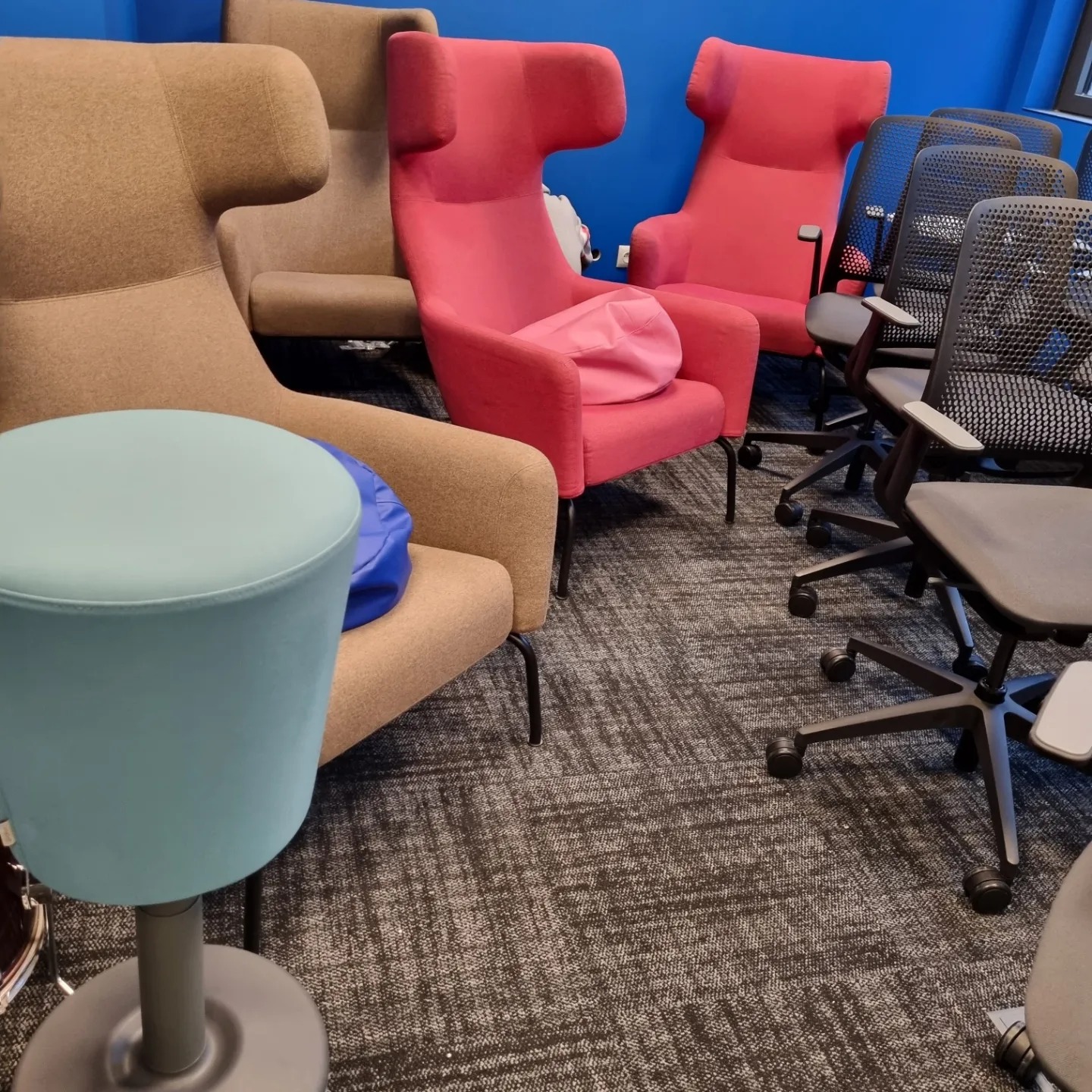 Różnokolorowe krzesła biurowe i stołek na kółkach ustawione na wzorzystej wykładzinie w biurze z niebieską ścianą w tle.