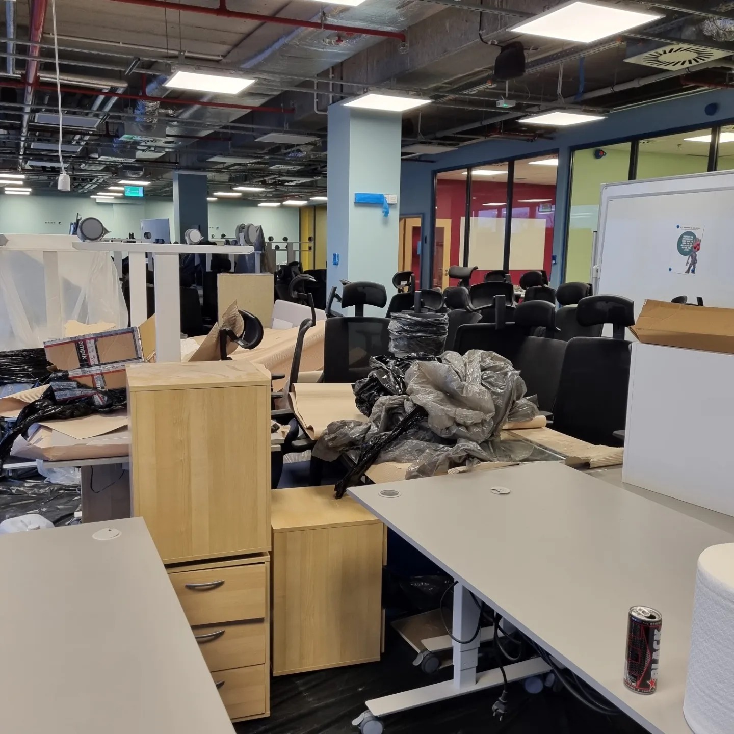 Biuro w trakcie porządkowania po remoncie, widoczne biurka, krzesła, kartony i worki na śmieci, puszka napoju energetycznego na blacie biurka.