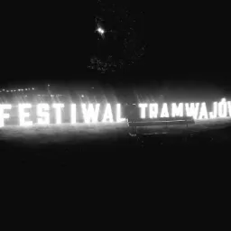 Oświetlony napis 'Festiwal Tramwajów' w nocy, ławka parkowa przed napisem, sylwetki ludzi w tle.