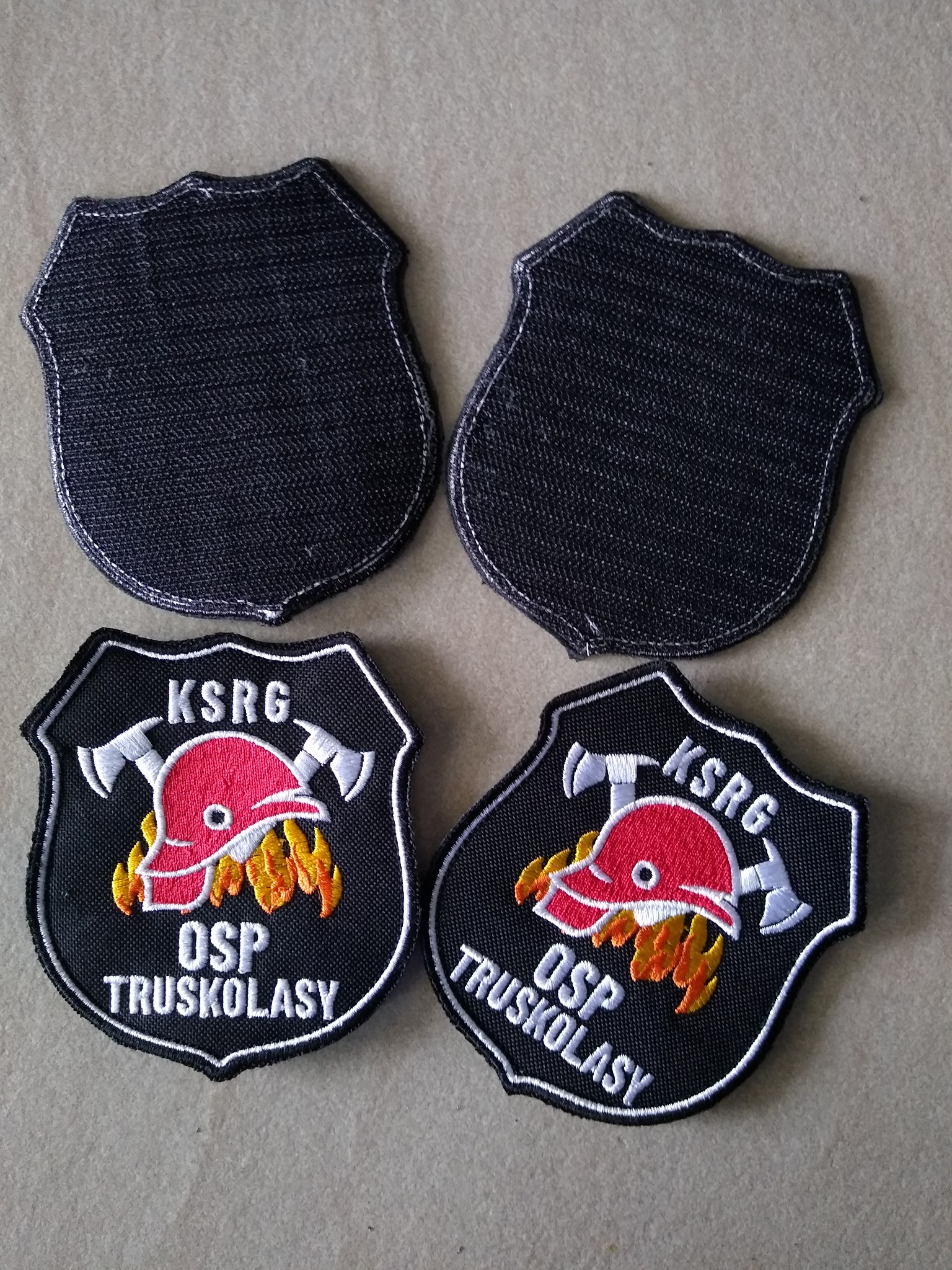 Cztery naszywki w kształcie tarczy: dwie czarne, puste i dwie z haftowanym logo OSP Truskolsy z hełmem strażackim i toporami na czarnym tle.