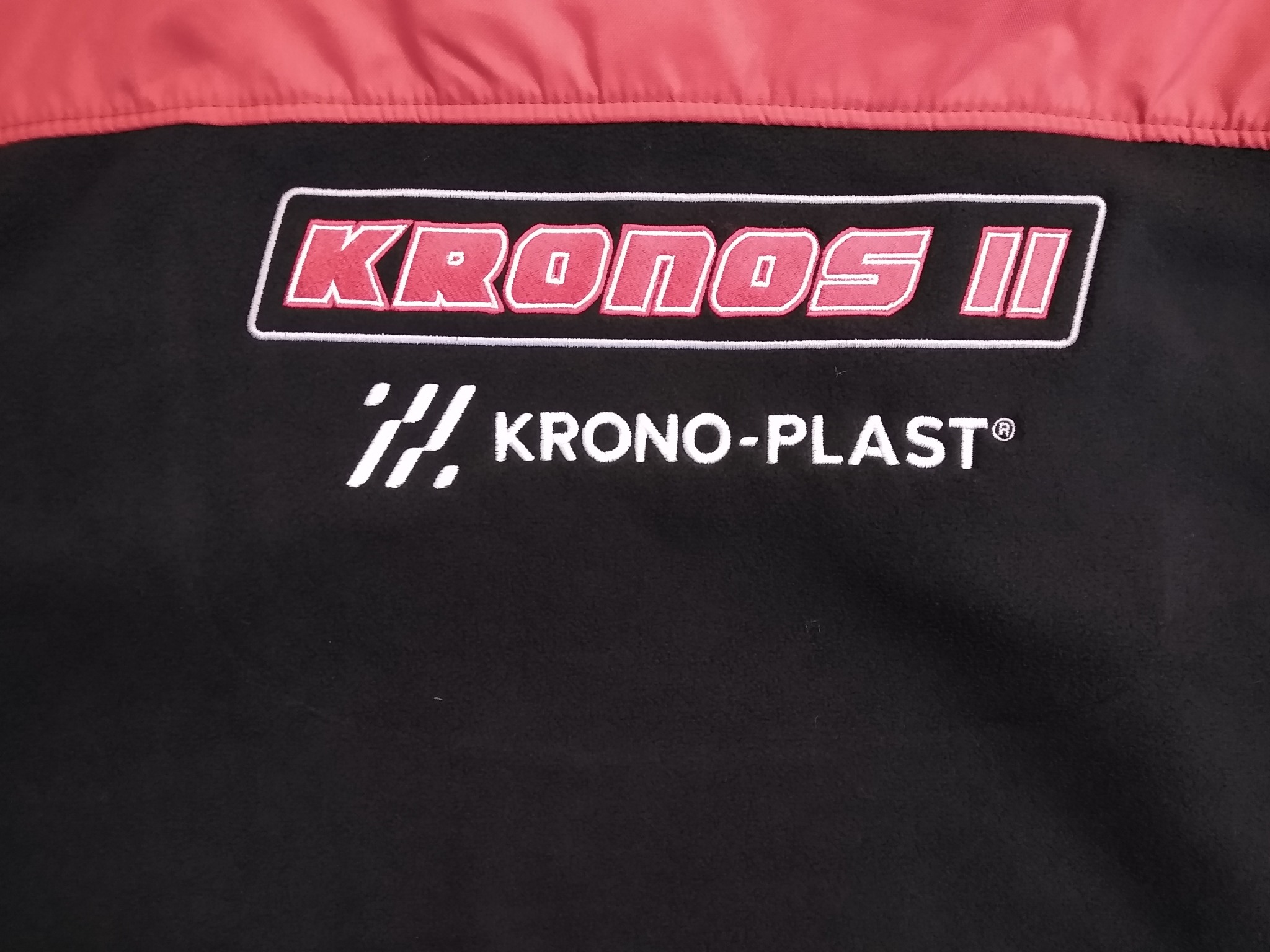 Część czarnej kurtki z czerwonym kołnierzem i wyhaftowanym logo 'KRONOS II' oraz 'KRONO-PLAST' poniżej.