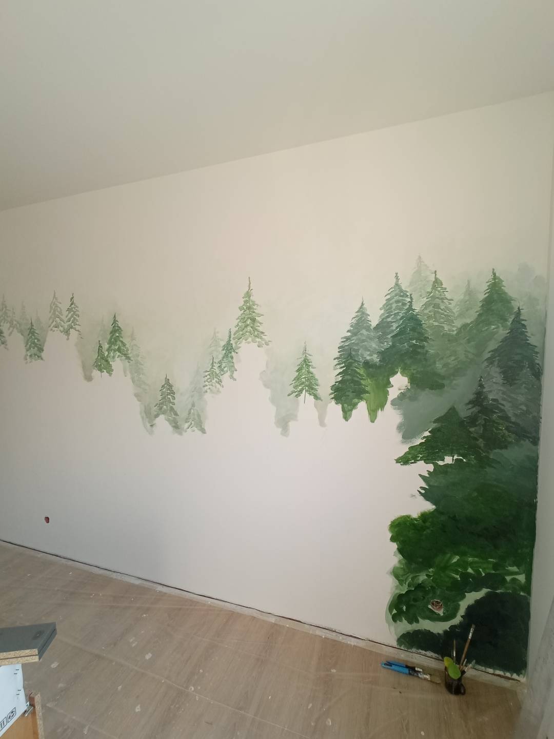 Ściana z ręcznie malowanym motywem leśnym, imitującym mglisty krajobraz z zielonymi drzewami iglastymi. Widoczne narzędzia malarskie.