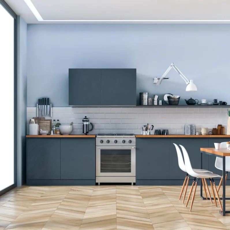 Nowoczesna kuchnia z szaro-niebieskimi szafkami, drewnianym blatem i podłogą w jodełkę. Białe krzesła i stół dopełniają aranżację. Ściana pomalowana na jasny odcień niebieskiego.