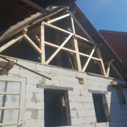 Wznoszenie drewnianej konstrukcji dachu z belek na murowanym budynku z otworami okiennymi, widok z dołu na tle błękitnego nieba.