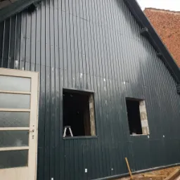 Ściana budynku obita blachą trapezową w kolorze grafitowym, z dwoma otworami okiennymi w trakcie montażu, częściowo otwarte drzwi wejściowe po lewej stronie.