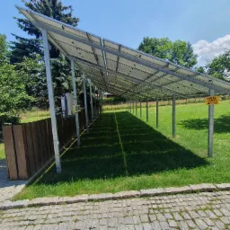 Instalacja fotowoltaiczna na metalowej konstrukcji w ogrodzie, widok z poziomu chodnika z kostki brukowej, tabliczka ostrzegawcza o spadającym lodzie i śniegu.