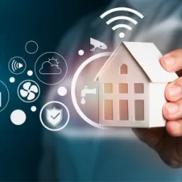 Dłoń trzyma model domu otoczonego ikonami: Wi-Fi, kamera monitoringu, głośnik, słońce za chmurą, kran, wentylator, tarcza z ptaszkiem, zamek, gniazdko z liściem. W tle cyfrowe tło z binarnym kodem.