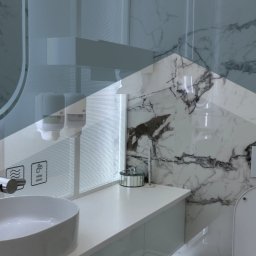 ASM Wroclaw Cleaning - Nowoczesna łazienka z marmurowymi płytkami, białą umywalką nablatową, dozownikami mydła i koszem na śmieci z pedałem; widoczny fragment toalety; logo firmy 'ASM Wrocław Cleaning' na górze.