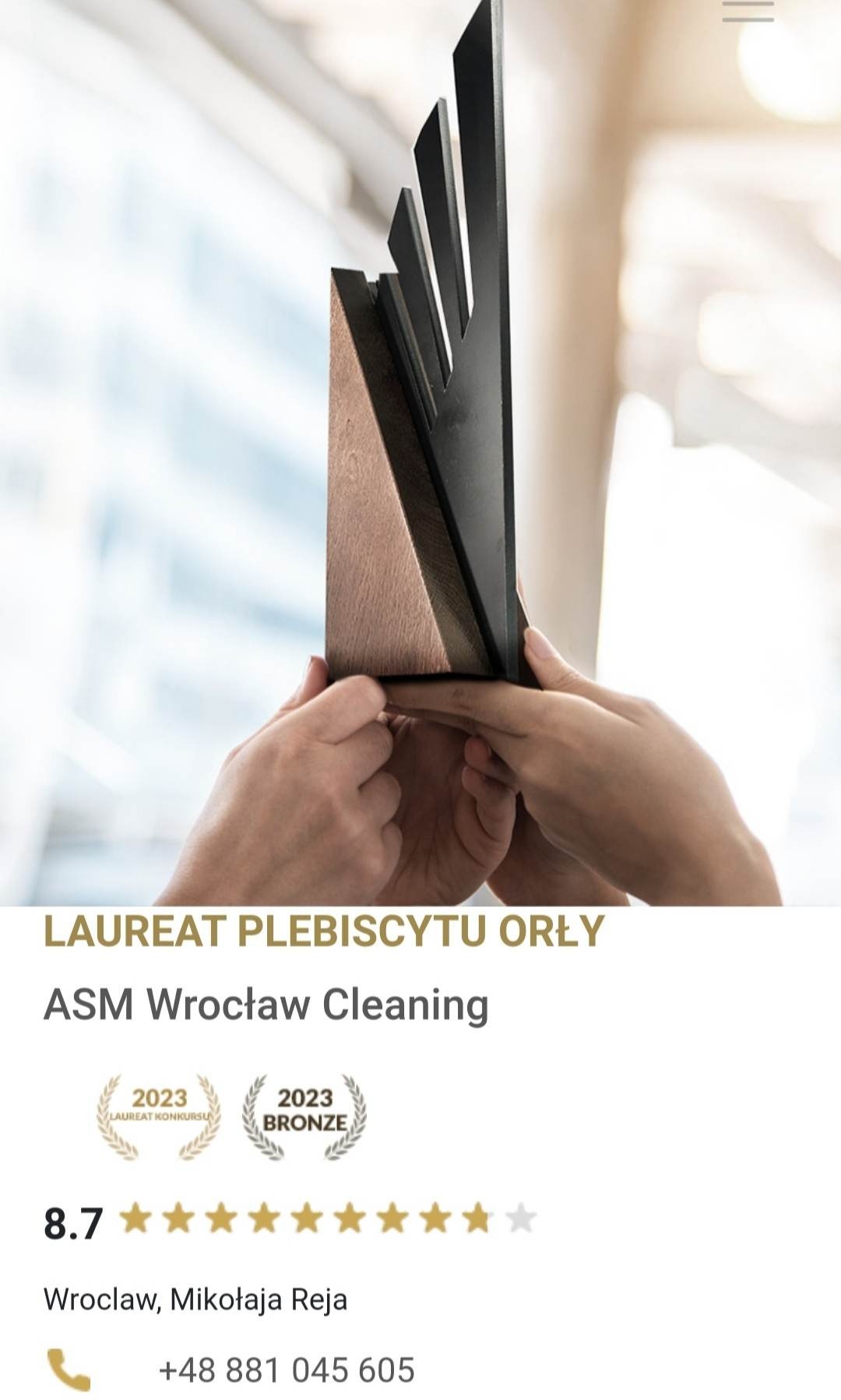 Ręce trzymające nowoczesną statuetkę z drewna i czarnego metalu, nagroda LAUREAT PLEBISCYTU ORŁY dla ASM Wrocław Cleaning, widoczne logo z rokiem 2023 i napisem LAUREAT KONKURSU oraz BRONZE.