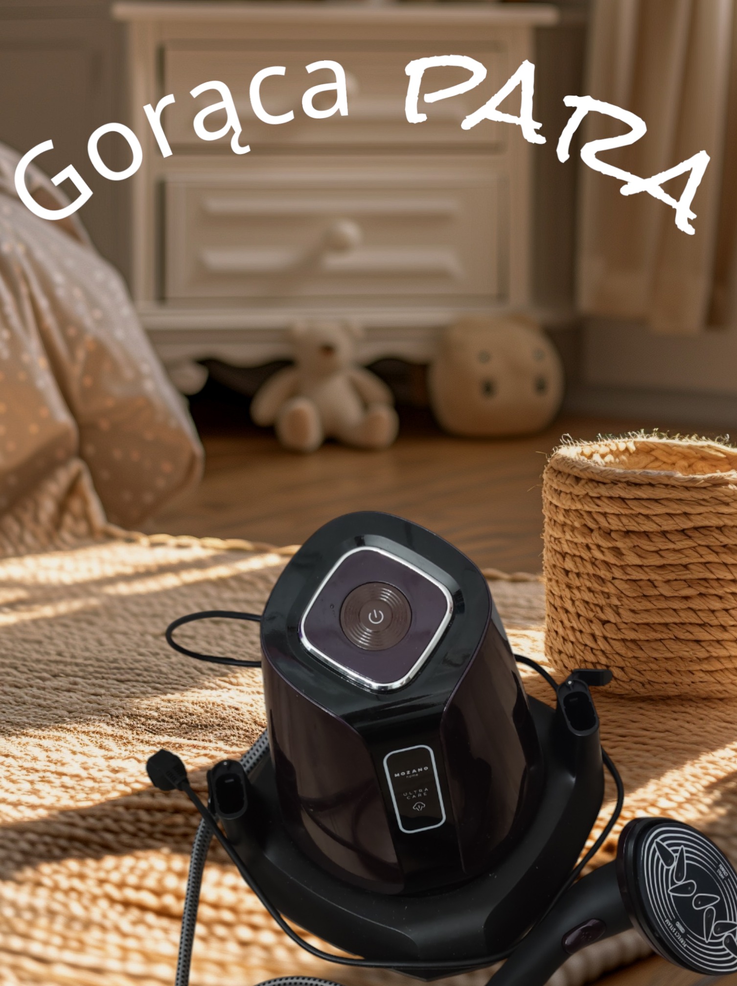 Parownica do ubrań marki Mozano Home Ultra Care na beżowym dywanie, w tle komoda i pluszaki, napis 'Gorąca PARA' nałożony graficznie.