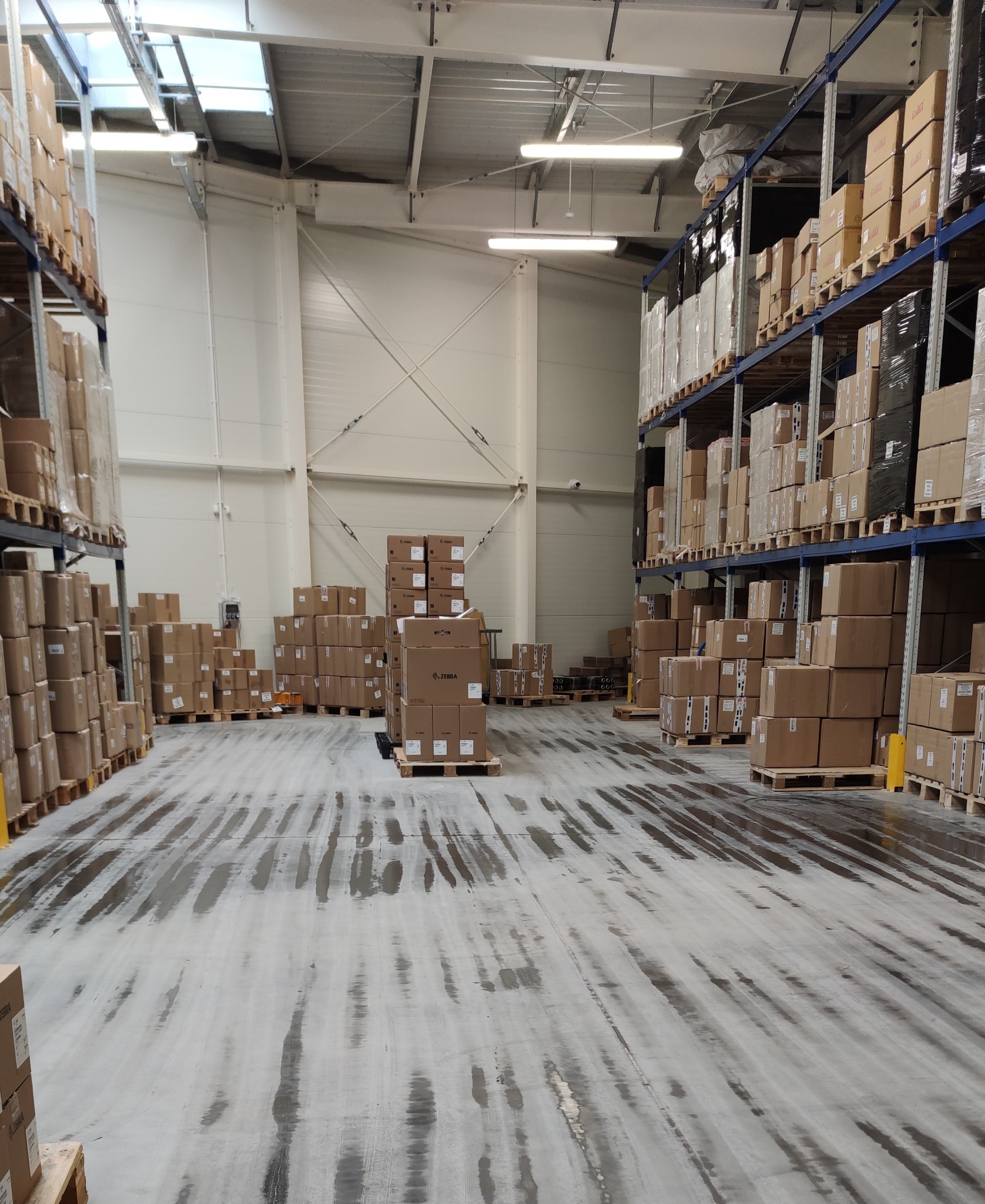 Wnętrze magazynu z regałami wypełnionymi kartonami, mokra betonowa podłoga z widocznymi śladami po myciu.