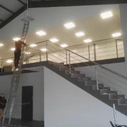 Montaż sufitu podwieszanego kasetonowego z oświetleniem LED, widoczny pracownik na drabinie i fragment schod&oacute;w z metalową balustradą.