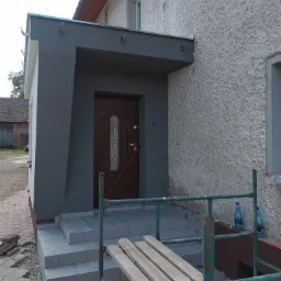 Wejście do domu z nową, szarą wiatrołapem i schodami wyłożonymi płytkami, widoczne rusztowanie i butelki z wodą, elewacja o grubej fakturze.