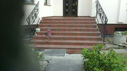 Schody wejściowe do domu w Nysie, wyłożone brązowymi płytkami, z czarnymi, kutymi balustradami, widoczne narzędzia pracy na schodach.