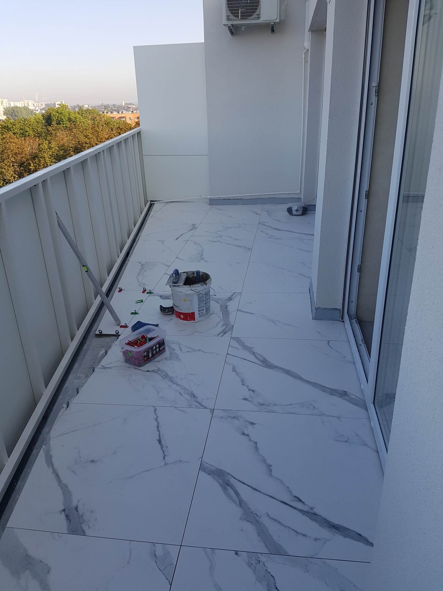 Balkon w trakcie układania białych płytek imitujących marmur, widoczne narzędzia glazurnicze, krzyżyki dystansowe i poziomica, z widokiem na miasto i drzewa.