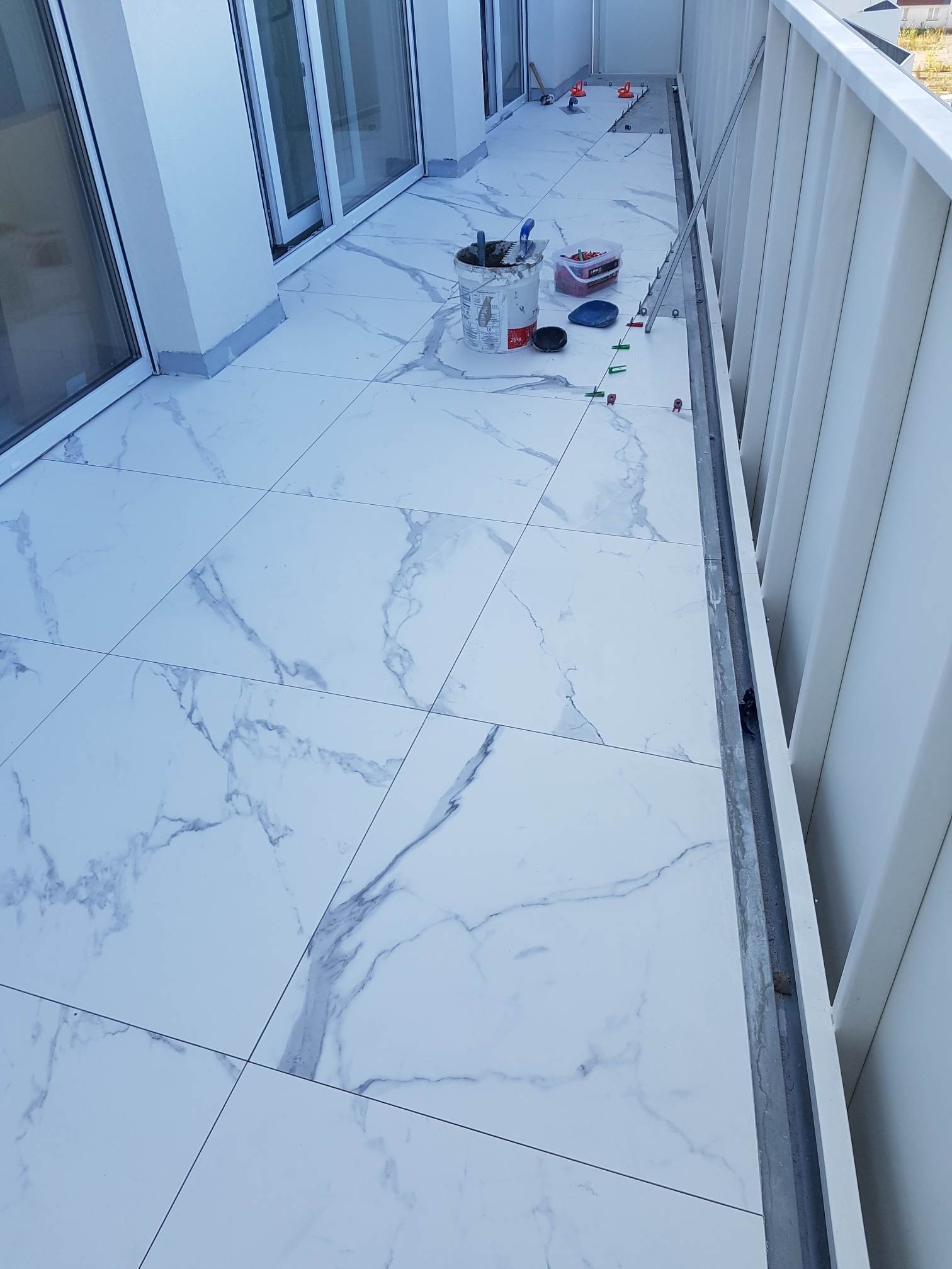 Białe płytki imitujące marmur ułożone na balkonie, widoczne narzędzia glazurnicze: wiadro, poziomica, krzyżyki dystansowe.