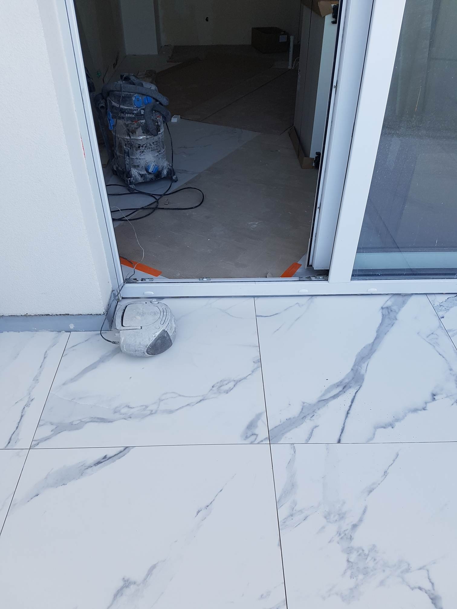 Białe płytki podłogowe imitujące marmur, z widocznym odkurzaczem przemysłowym i radiem budowlanym, sugerujące prace wykończeniowe w pomieszczeniu.