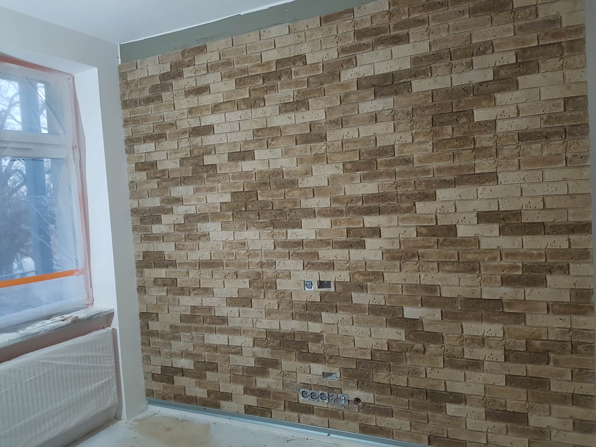 Ściana wewnętrzna wyłożona dekoracyjną cegłą w odcieniach beżu i brązu, z widocznymi puszkami elektrycznymi i zabezpieczonym folią oknem.