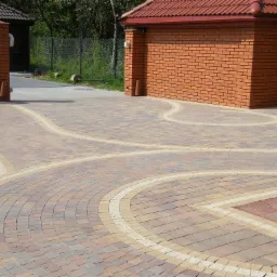 Wzorzysta nawierzchnia z kostki brukowej w odcieniach brązu i beżu, z białymi liniami oddzielającymi wzory, prowadząca do ceglanego budynku z czerwonym dachem i słupków z cegły.