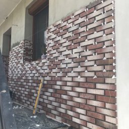 Good Value - Montaż cegieł klinkierowych na elewacji budynku z użyciem krzyżyków dystansowych, widoczny poziomica i częściowo zakryte folią podłoże.