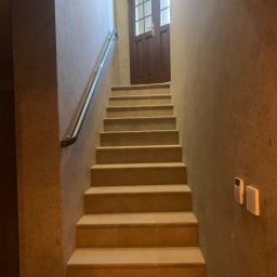 Good Value - Klatka schodowa z beżowymi płytkami, metalowa poręcz po lewej stronie i ciemne, przeszklone drzwi na końcu schodów, ściany o nierównej fakturze w odcieniach beżu i brązu, widoczne panele sterowania...