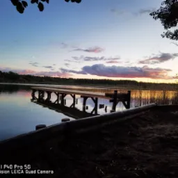 Pomost drewniany nad jeziorem o zachodzie słońca, widoczne odbicie konstrukcji w wodzie i trzciny na drugim brzegu.