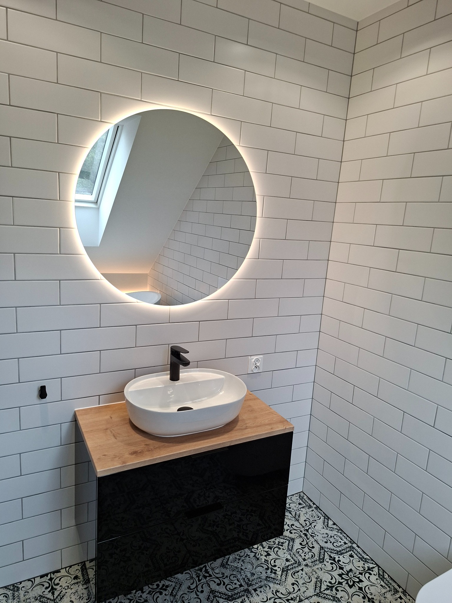 Biała łazienka z umywalką nablatową, okrągłym lustrem z podświetleniem LED i płytkami imitującymi cegłę. Czarna szafka pod umywalką i wzorzysta podłoga.
