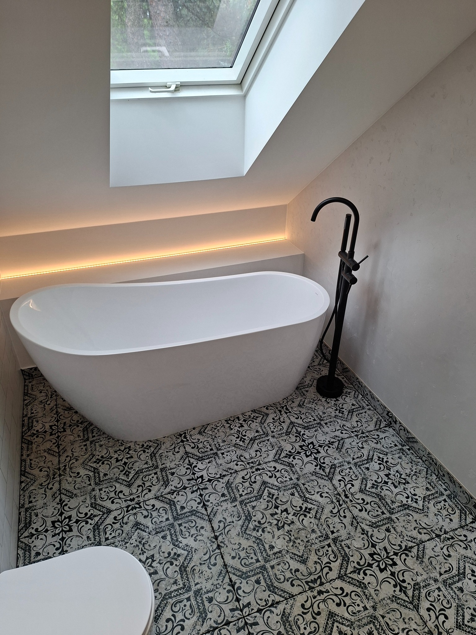 Nowoczesna łazienka na poddaszu z wolnostojącą wanną, czarną armaturą i wzorzystą podłogą. Okno dachowe doświetla wnętrze, a listwa LED dodaje ciepłego światła.