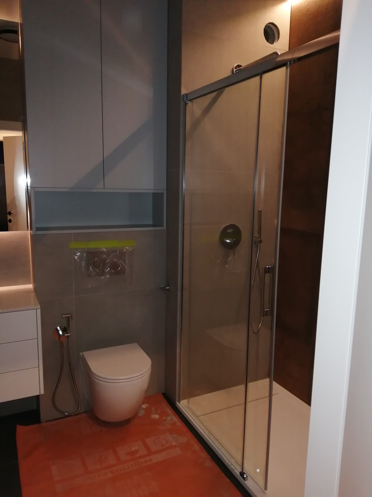 Nowoczesna łazienka z kabiną prysznicową typu walk-in, wiszącą toaletą i szarą zabudową meblową. Podłoga zabezpieczona folią ochronną.
