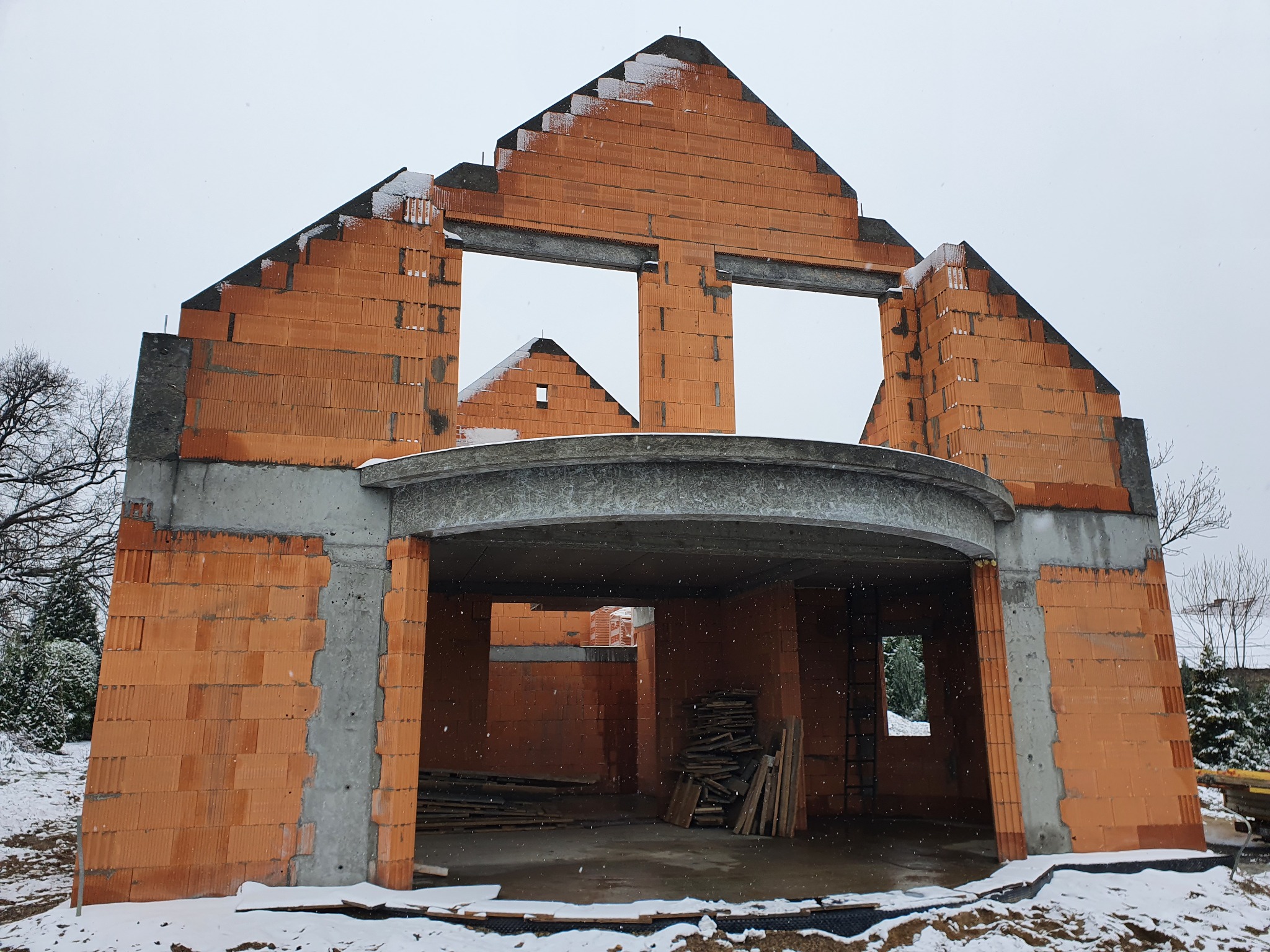 Surowy stan domu z czerwonej cegły ceramicznej, widoczny w zimowej scenerii, z elementami konstrukcyjnymi pokrytymi śniegiem, w tle drzewa bez liści.