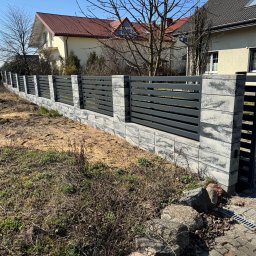 Nowoczesne ogrodzenie panelowe z szarego betonu i grafitowych przęseł, widoczne z bliska na tle domów i błękitnego nieba. Elementy betonowe z wyraźną fakturą.