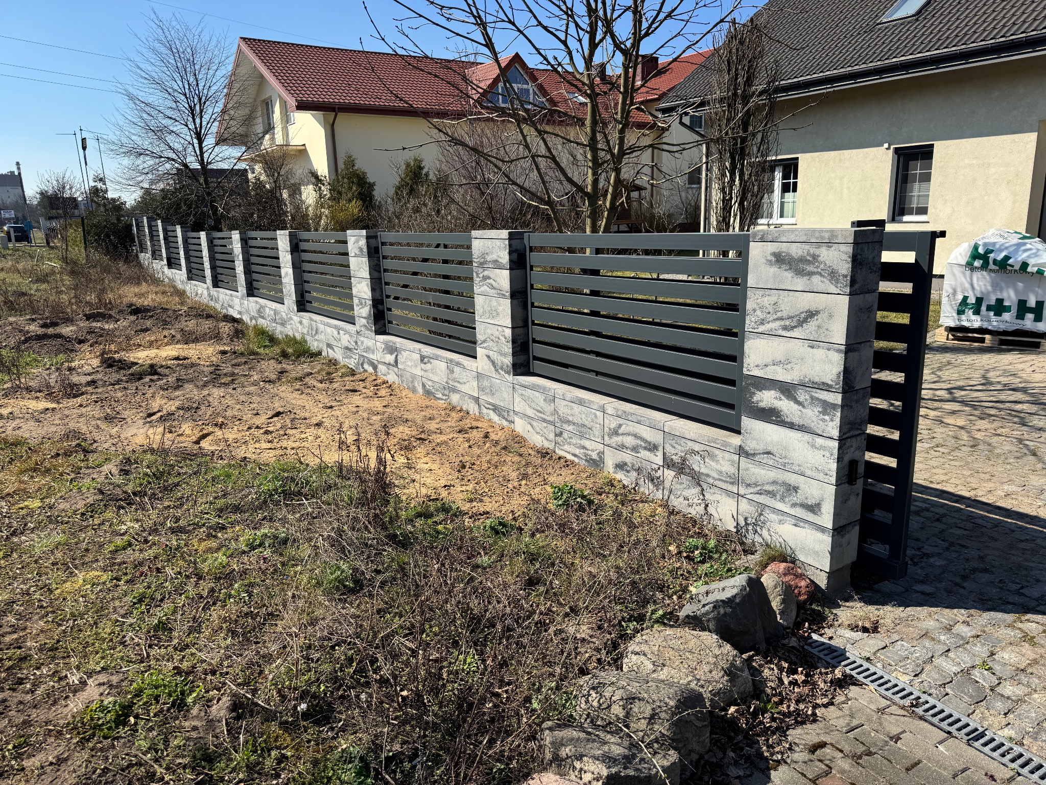 Nowoczesne ogrodzenie panelowe z szarego betonu i grafitowych przęseł, widoczne z bliska na tle domów i błękitnego nieba. Elementy betonowe z wyraźną fakturą.