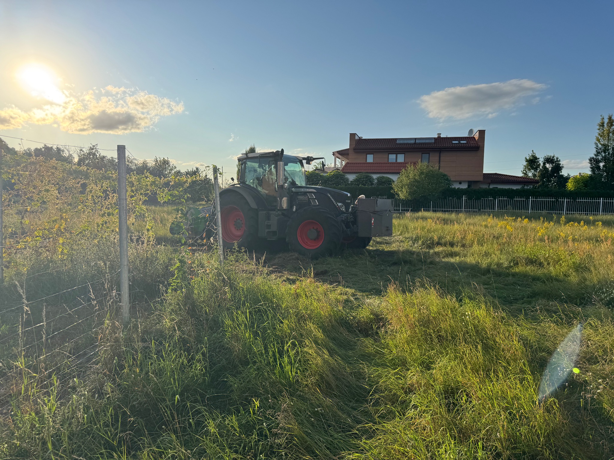Czarny traktor Fendt z czerwonymi felgami na polu z częściowo skoszoną trawą, w tle dom z brązową elewacją i błękitne niebo z chmurami.