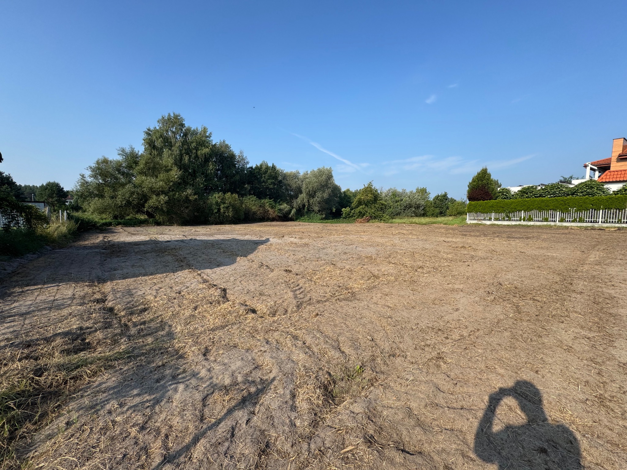 Przygotowany teren pod budowę z widocznymi śladami opon, otoczony zielenią i ogrodzeniem. Błękitne niebo w tle. Cień osoby robiącej zdjęcie w prawym dolnym rogu.