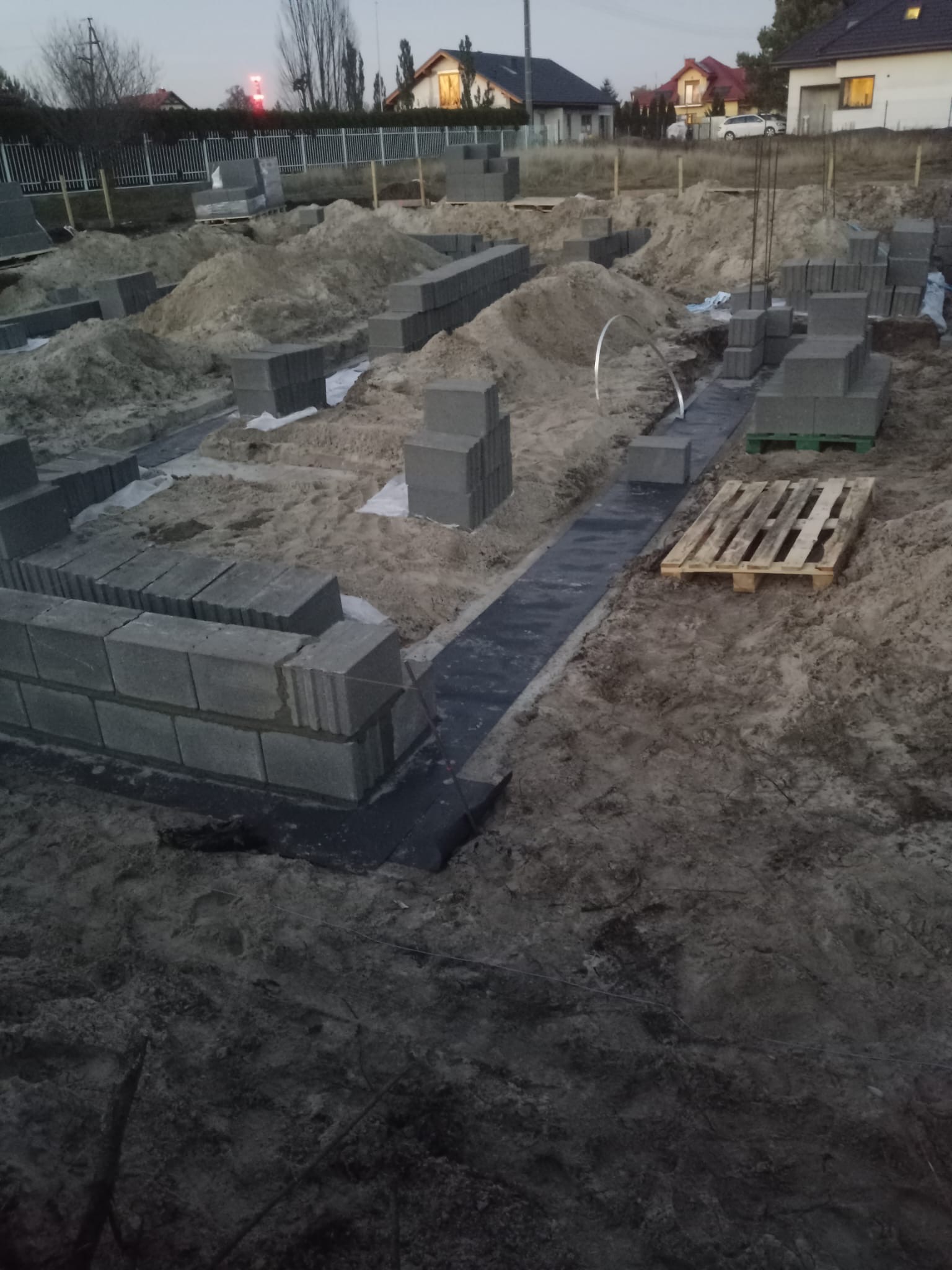 Wykop pod fundamenty domu z ułożonymi bloczkami betonowymi i izolacją przeciwwilgociową na piasku. Widoczne palety i domy w tle.
