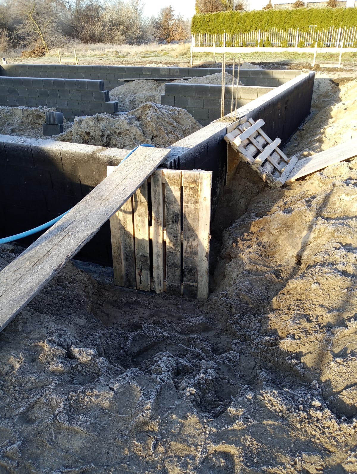 Fundamenty domu w trakcie budowy, zabezpieczone deskami i paletami. Widoczne bloczki fundamentowe, izolacja i wykop. Ziemia pokryta szronem.