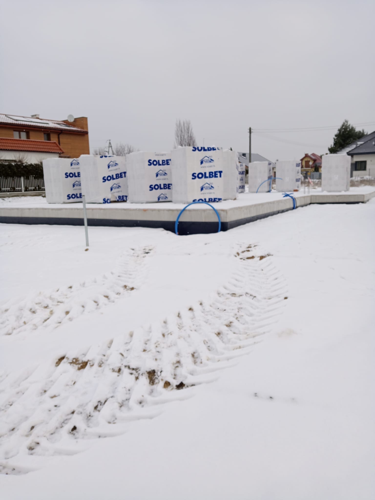 Fundament domu w zimowej scenerii. Na fundamencie ułożone bloczki SOLBET, zabezpieczone niebieskim sznurkiem. Ślady opon na śniegu. W tle zabudowa jednorodzinna.