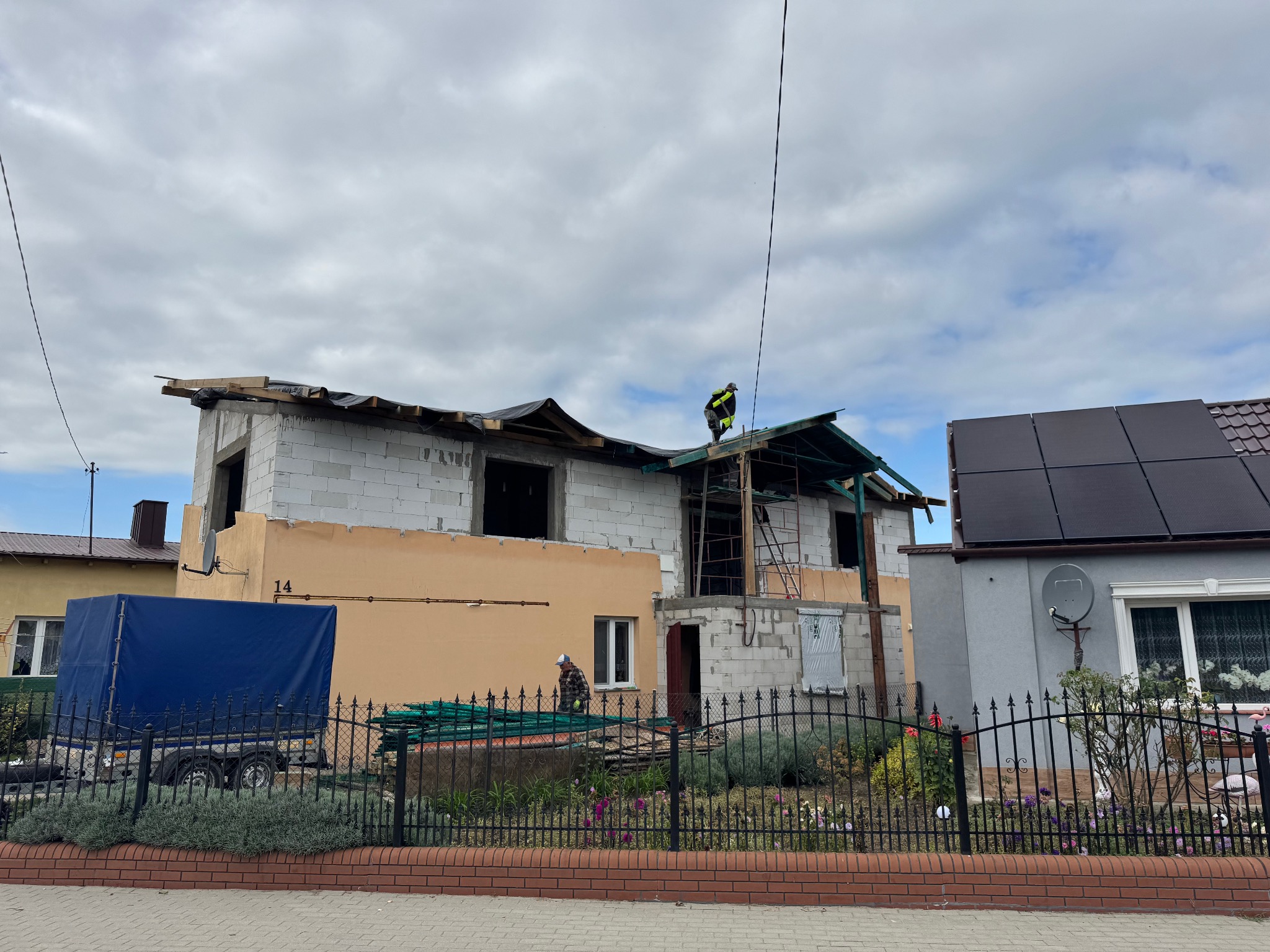 Budowa domu w trakcie realizacji: widoczne ściany z pustaków, częściowo ocieplona elewacja, pracownik na dachu. W tle dom z panelami słonecznymi i przyczepa.